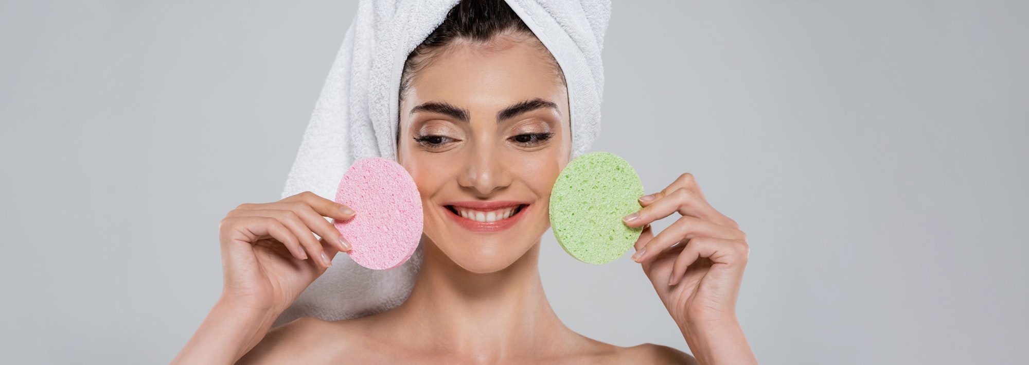 happy young woman with towel on head holding beauty sponges isolated on grey, banner